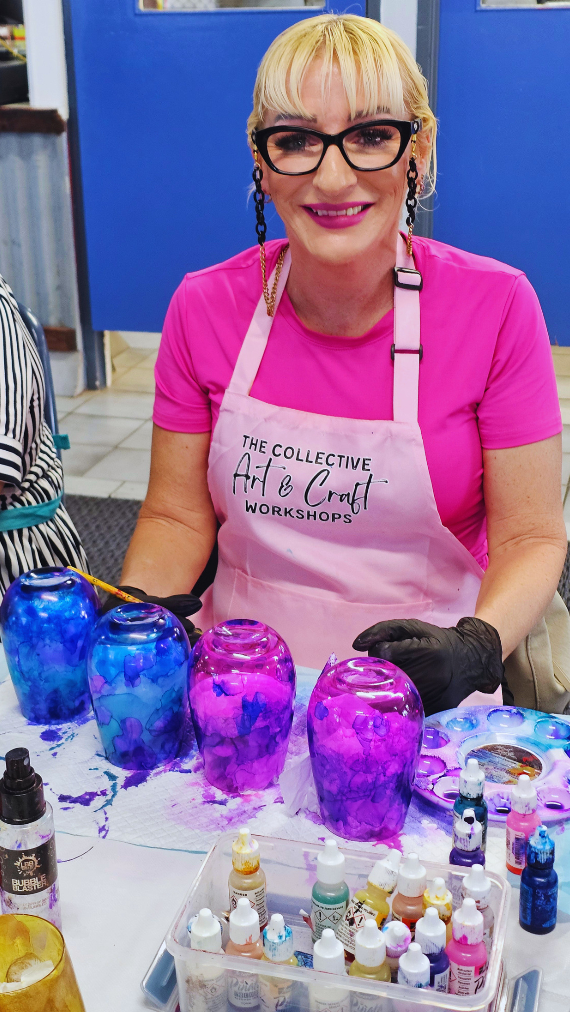 A woman wearing a pink apron that reads 'The Collective Art & Craft Workshops' paints four glass jars with vibrant blue and purple marbled paint. She is wearing a bright pink t-shirt, black glasses, and black gloves, with various paint bottles and a palette on the table in front of her.
