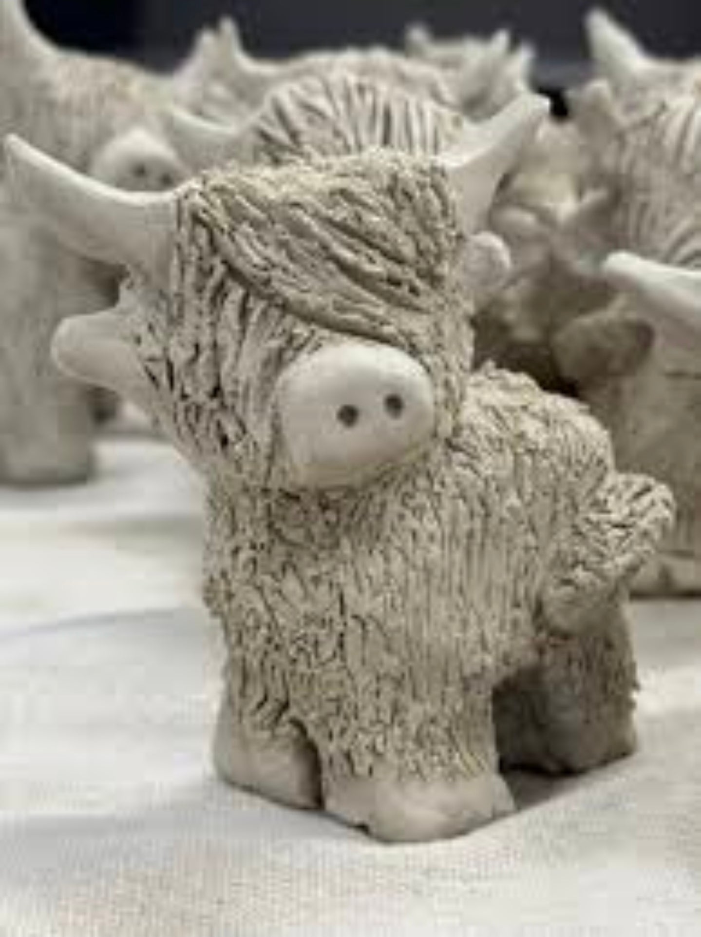 A small Highland cow figurine with textured fur, horns, and a pinkish snout, shown in a light gray clay or resin finish on a white surface with other similar figurines blurred in the background.