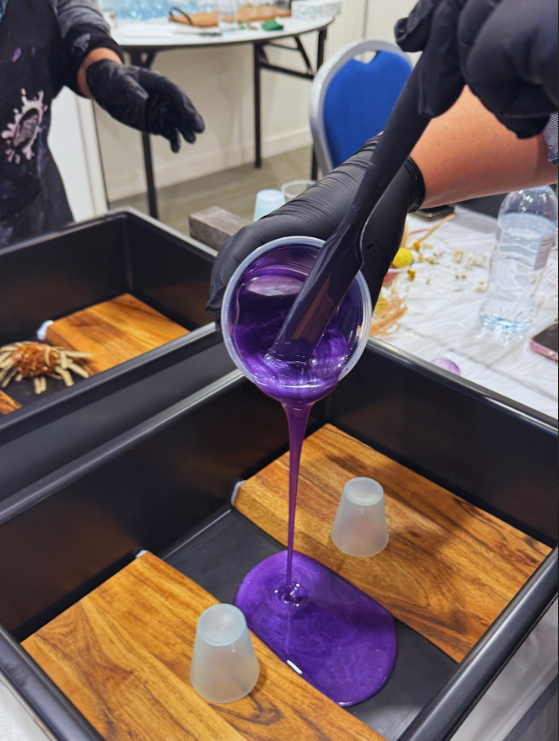 Person pouring purple liquid into a mold on a wooden surface with gloves on.